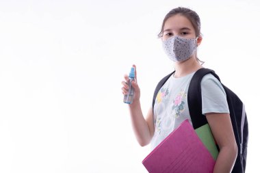 Yüzünde gri bir maske olan okul kızı elinde beyaz bir arka planda antiseptik ve defter tutuyor.