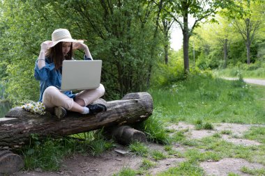 Kırsalda, şapkalı genç bir kız eski bir kütüğün üzerinde oturuyor, elinde dizüstü bilgisayarla, baharda.