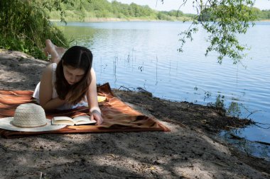 Yazın elinde kitapla güzel bir gölün kıyısında dinlenen beyaz elbiseli bir kız.