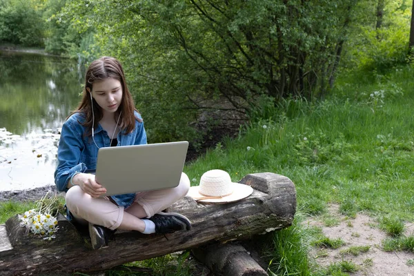 in the countryside, near the lake, a young girl sits on an old log with a laptop, in spring