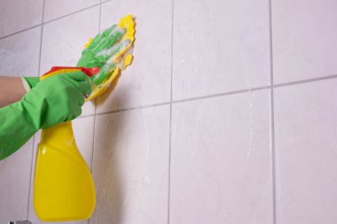Kız banyoyu temizliyor ve beyaz arka planda deterjanla fayanslardan bir duvar yıkıyor.
