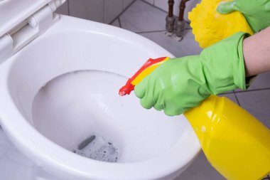 Banyodaki temizlikçi kadın tuvaleti yeşil eldivenlerle ve beyaz arka planda deterjanla yıkıyor.
