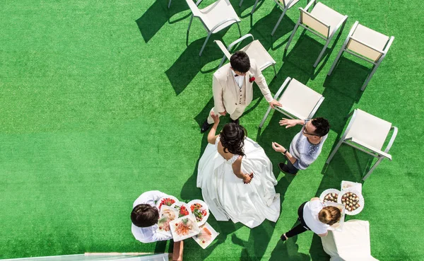 La vista superior del novio y la novia hablando con el organizador contra el telón de fondo de sillas blancas y hierba verde y los camareros que distribuyen la comida . — Foto de Stock