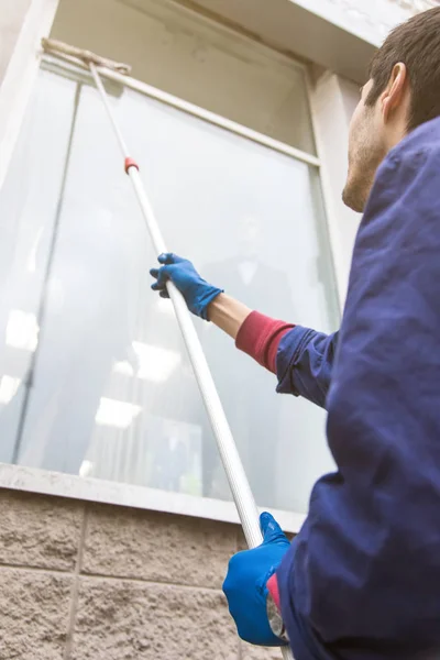 Un joven limpiador de monos azules lava las ventanas y el frente de la tienda. Servicio de limpieza — Foto de Stock