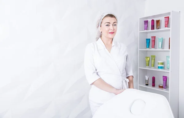 A beautician woman in a white coat is standing near a chair on the background of shelves with creams. Profession of cosmetology and skin care — Stock Photo, Image