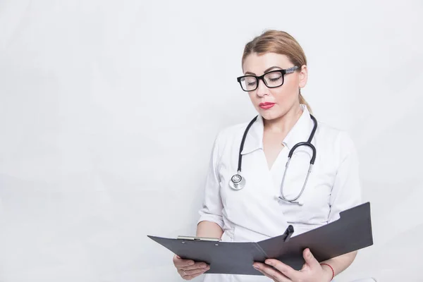 Retrato de uma médica com óculos em um vestido médico branco com um estetoscópio segurando uma pasta preta com notas de pacientes em um fundo leve — Fotografia de Stock