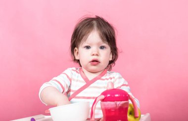 Parlak renkli hafif pembe bir arka plan üzerinde bir çocuğun sandalyede oturan küçük bir kız bebek yiyor