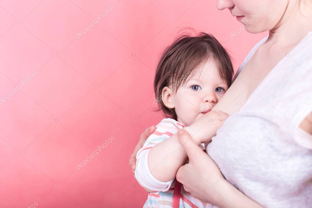 Little girl sucks mother's breast on bright colored light pink background