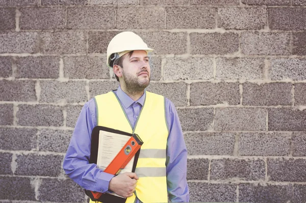 A young male engineer in a white helmet, safety glasses and a yellow vest against a wall of concrete blocks holds a folder of documents and instruments