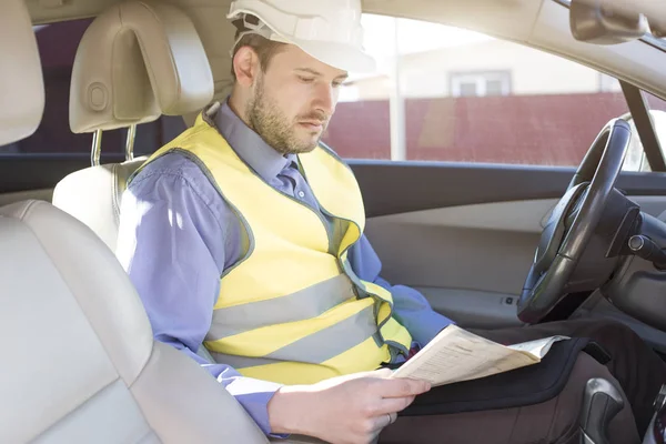 Man engineer builder in a white hard hat, shirt and yellow waistcoat sits in the car and reads