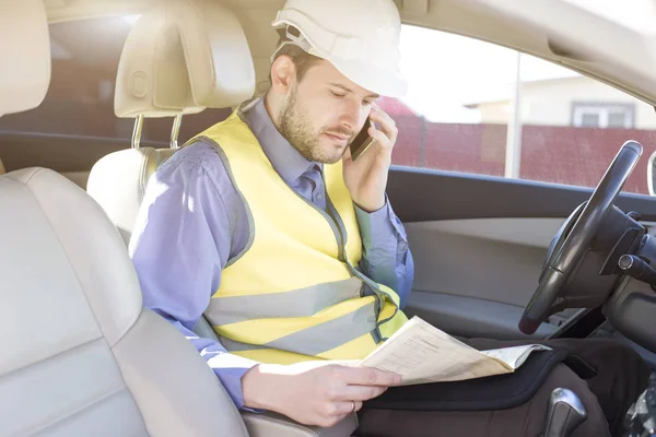 Engenheiro de construção masculino em capacete branco, camisa e colete amarelo senta-se no carro e digitaliza documentos enquanto fala ao telefone — Fotografia de Stock