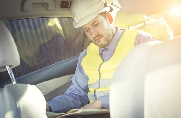 Man engineer builder in a white hard hat, shirt and yellow waistcoat sits in the car and reads