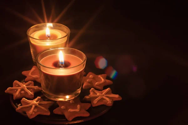 Burning candles in transparent glasses and gingerbread cookie on a saucer on a dark background. mockup for postcards or greetings with copy space — Stock Photo, Image