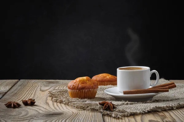 Hot coffee with steam in a white cup with muffins, cinnamon and star anise on burlap on a wooden table on a black concrete background — Stock Photo, Image