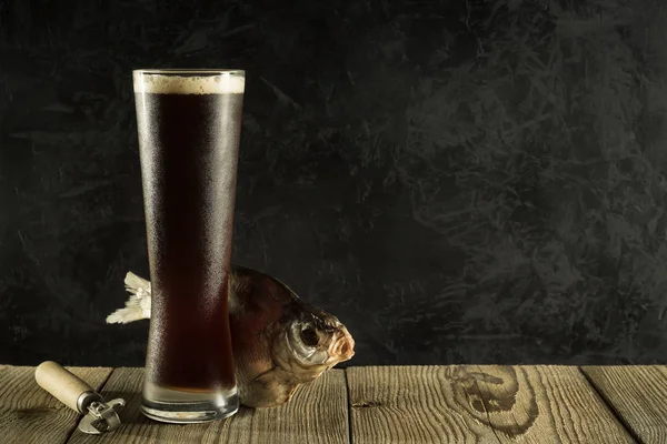 Vaso lleno de cerveza espumosa fría oscura con pescado de río seco en tablas ásperas contra una pared de hormigón negro. artista maqueta creativa con espacio de copia. vista lateral Fotos de stock libres de derechos