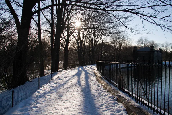 Nieve en la pasarela junto al lago y valla en el parque con siluetas — Foto de Stock