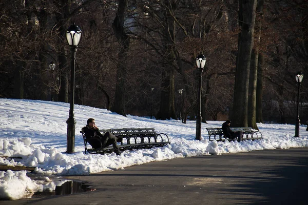 Central Park, Manhattan, Nueva York, 17 de marzo de 2017: la gente se sienta en los bancos del parque junto al poste de luz con nieve — Foto de Stock