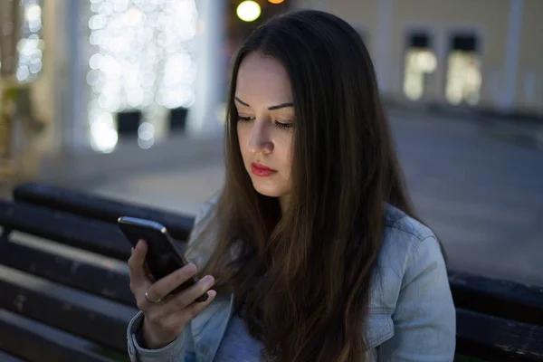 Mujer usando teléfono inteligente sentado en el banco en la ciudad de la noche — Foto de Stock