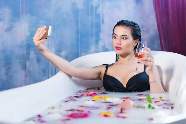 Sexy brunette woman taking selfie having a hot milk bath with flowers — Stock Photo, Image