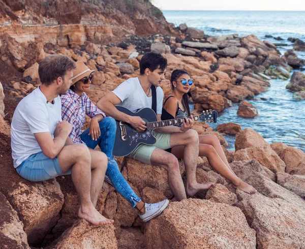 Amici seduti su pietre sulla spiaggia. man sta suonando la chitarra . — Foto Stock
