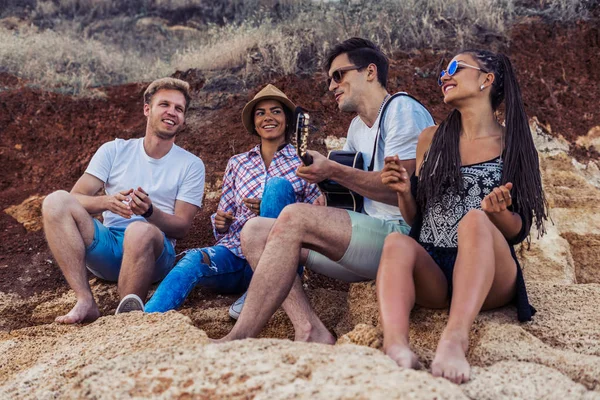 Amigos sentados em pedras na praia. homem está tocando guitarra . — Fotografia de Stock