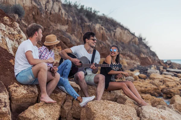 Amigos sentados em pedras na praia. homem está tocando guitarra . — Fotografia de Stock