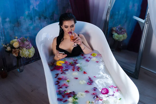 Sexy brunette woman is sitting in bath and chatting on smartphone — Stock Photo, Image