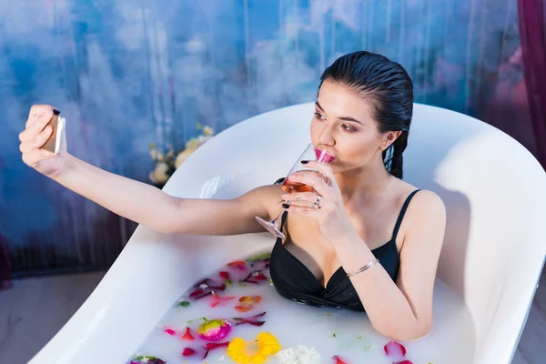 Sexy brunette woman taking selfie having a hot milk bath with flowers — Stock Photo, Image