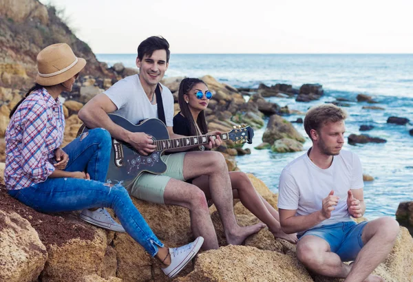 Amigos sentados en piedras en la playa. hombre está tocando la guitarra . — Foto de Stock