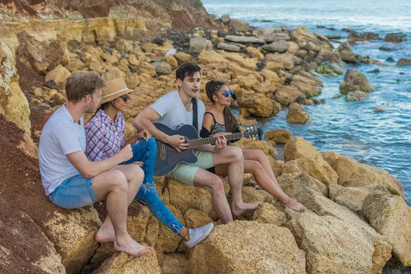 Amici seduti su pietre sulla spiaggia. man sta suonando la chitarra . — Foto Stock