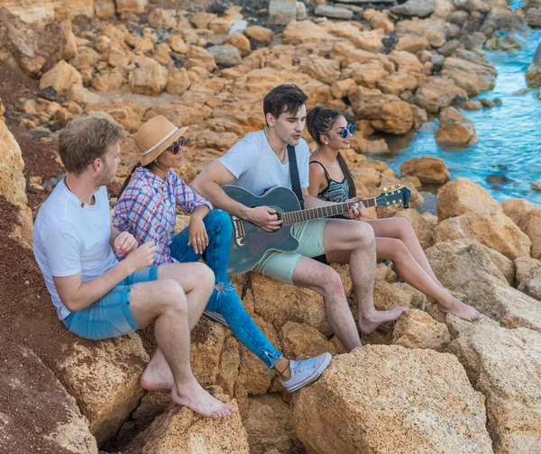 Przyjaciółmi siedząc na kamienie na plaży. człowiek jest gra na gitarze. — Zdjęcie stockowe