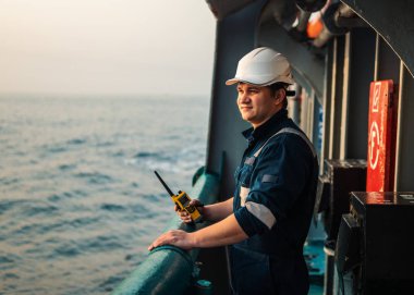 Marine Deck Officer or Chief mate on deck of offshore vessel or ship , wearing PPE personal protective equipment - helmet, coverall. He holds VHF walkie-talkie radio in hands. clipart