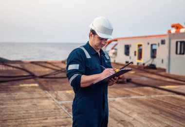 Güverte görevlisi offshore gemi veya gemi güvertede, PPE kişisel koruyucu ekipman giyiyor. Kontrol listesini doldurur. Denizdeki evraklar