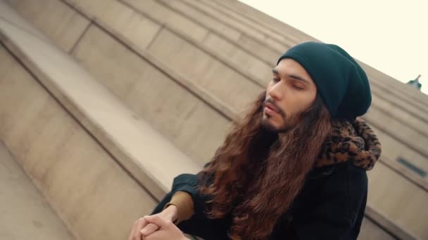 Portrait of long haired hipster sitting on street in modern city — Stock Video