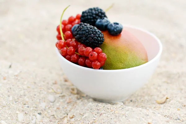 Summer fruits in a bowl on a background of sea sand. Summer vacation on the beach. Ripe whole  mango and berries of blackberry, raspberry, red currant in a bowl. Breakfast in the sand