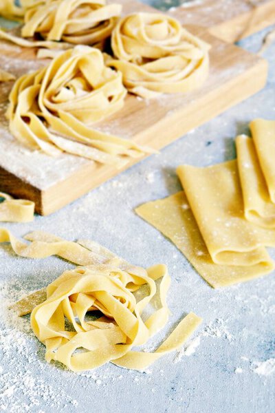 Homemade italian pasta, ravioli, fettuccine, tagliatelle on a wooden board and on a blue background. The cooking process, raw pasta. Tasty raw ravioli with ricotta and spinach,with flour on background