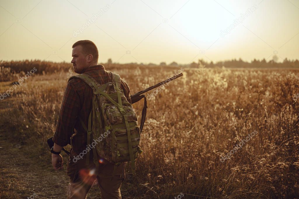 Humans back in checkered shirt with military backpack and shotgun in his hand, in the sun light. It's  a soldier hiding into the fields, or a hunter, walking in the grass and searching for prey