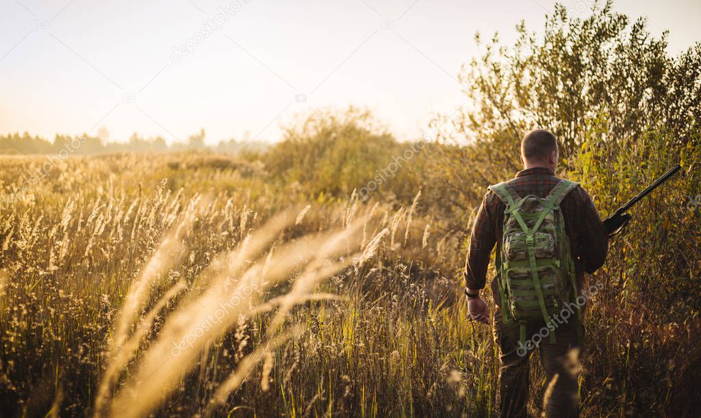 Humans back in checkered shirt with military backpack and shotgun in his hand, in the sun light. It's  a soldier hiding into the fields, or a hunter, walking in the grass and searching for prey