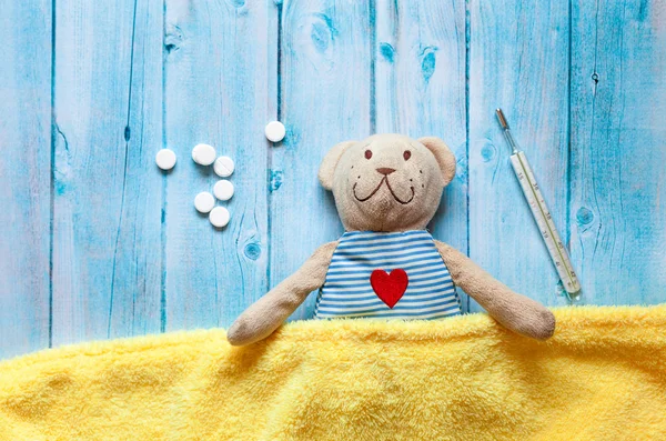 Crianças urso de pelúcia de brinquedo macio na cama com termômetro e pílulas, tomar a temperatura de um copo de mercúrio. Em fundo de madeira azul cobertor amarelo. Brincando no hospital . — Fotografia de Stock