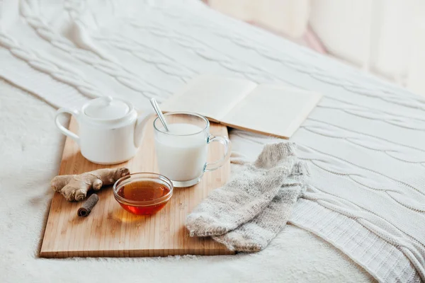 Leche caliente en una taza de vidrio y miel en una tabla de madera. El tratamiento de los niños una bebida caliente. Tratamiento de remedios caseros en la cama. Calcetines de punto caliente. El libro para leer . — Foto de Stock