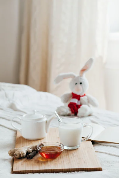 Du lait chaud dans une tasse en verre et du miel sur une planche en bois. Traitement des enfants une boisson chaude. Traitement des remèdes populaires au lit. Lapin en peluche pour votre enfant . — Photo