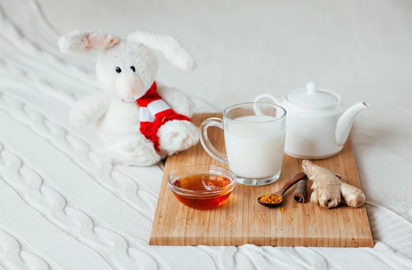 Leche caliente en una taza de vidrio y miel en una tabla de madera. El tratamiento de los niños una bebida caliente. Tratamiento de remedios caseros en la cama. Conejito de juguete suave para su hijo . — Foto de Stock