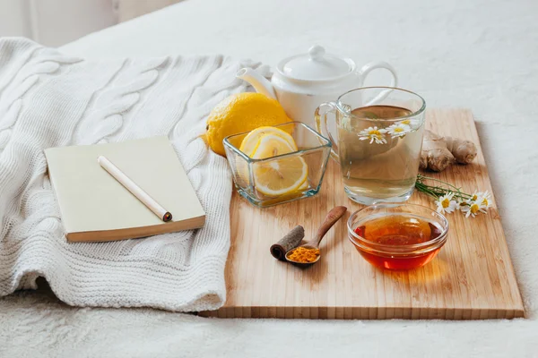 Té de hierbas con flores de manzanilla, cúrcuma y miel en una tabla de madera. Tratamiento de la bebida caliente con jengibre. Tratamiento de remedios caseros en la cama. Ocio con libro . — Foto de Stock