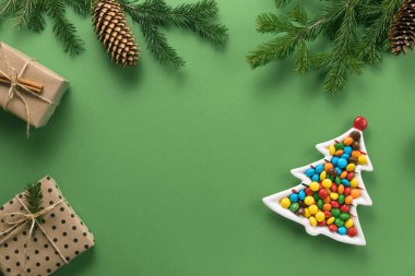 Noel kompozisyonu. Ladin 'den hediyeler ve dallar. Yeşil arka planda renkli şekerlerle dolu bir Noel ağacı şeklinde bir tabak. Düz konum, üst görünüm, kopyalama alanı