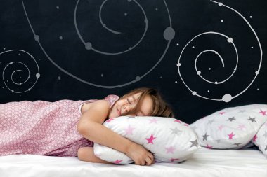 Anaokulu çağındaki sevimli küçük kız yıldızlı bir yastıkta uyuyor. Uyku vakti. Uzay ve bir çocuğun hayalleri hakkında soyut bir arka plan. Rüyada bir yolculuk. Mutlu çocukluk.