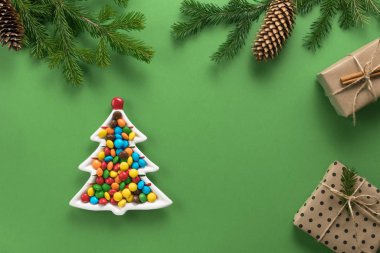 Noel kompozisyonu. Ladin 'den hediyeler ve dallar. Yeşil arka planda renkli şekerlerle dolu bir Noel ağacı şeklinde bir tabak. Düz konum, üst görünüm, kopyalama alanı