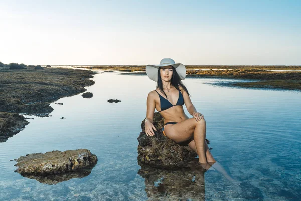 Retrato de estilo de vida de verano de una mujer bronceada bastante joven. Disfrutando de la vida y sentado en la playa de la isla tropical. Con un bikini elegante y un sombrero de ala ancha con rayas. Mirando al mar —  Fotos de Stock