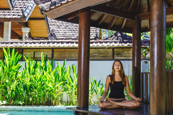 Female meditating by the pool, Bali, Indonesia.The girl sits in a gazebo at a bali in a lotus pose and meditates with a view of the pool.