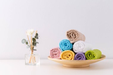 Rafta yuvarlanmış renkli banyo havluları açık gri arka planda.