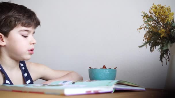 Чарівний 6 років хлопчик читає книгу і їсть сухофрукти — стокове відео